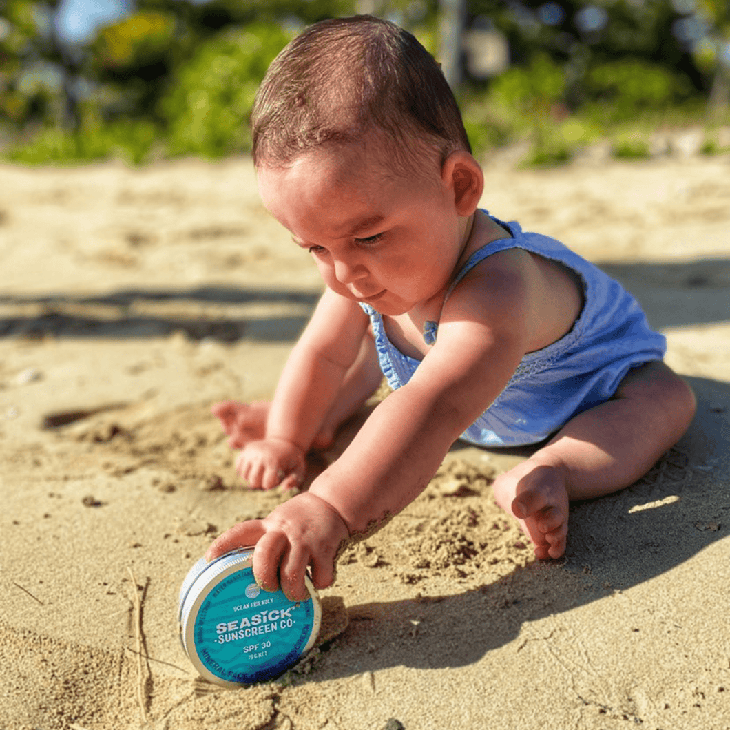 baby rolling a tin of sunscreen on a sandy beach