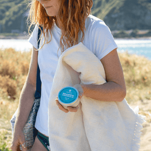 woman with orange hair at the beach carrying a beach bag blanket and tin of sunscreen