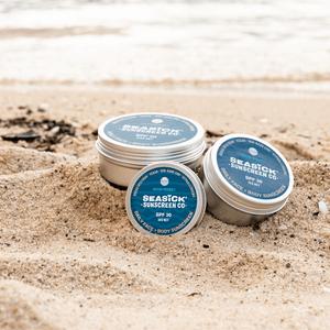 three silver tins of sunscreen of varying sizes siting on sand with ocean in background